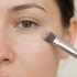 Brush gently blending concealer under the eye, minimal product, clean skin, soft lighting.