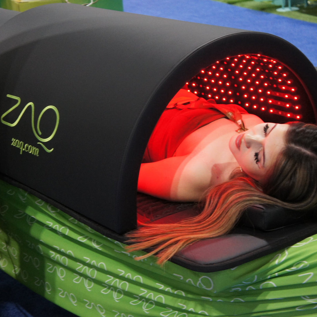 Person lying inside a ZAQ device with red light therapy on a green blanket.
