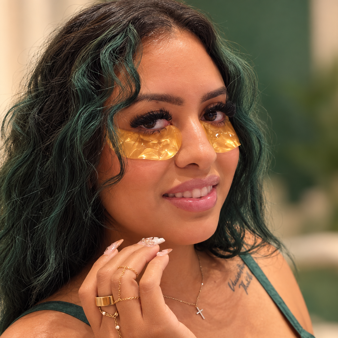 A close-up lifestyle shot of a woman with dark green highlighted hair wearing gold eye patches, a green top, and gold jewelry against a softly blurred background.