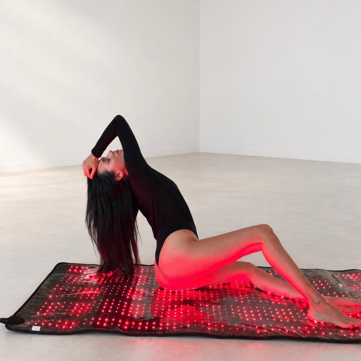 A woman in a black bodysuit reclines on a red LED light therapy mat in a minimalist white studio setting.