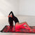 A woman in a black bodysuit reclines on a red LED light therapy mat in a minimalist white studio setting.