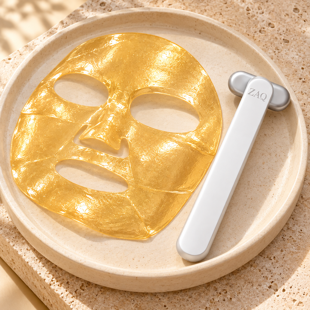 A gold facial sheet mask and a white handheld roller tool with a silver handle are arranged on a round beige tray. The roller has the brand name ZAQ printed on it.