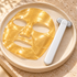 A gold facial sheet mask and a white handheld roller tool with a silver handle are arranged on a round beige tray. The roller has the brand name ZAQ printed on it.