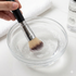 Makeup brush being cleaned in a bowl of soapy water with a bottle of cleaning solution in the background.