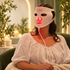 A woman wearing a white ZAQ LED light therapy face mask that glows red on the nose and mouth areas, sitting in a cozy, warmly lit room.