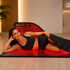 A woman lies on a red LED light therapy mat in a modern spa-like room, wearing a black sports bra and leggings. The mat has a black frame with a red illuminated grid pattern.